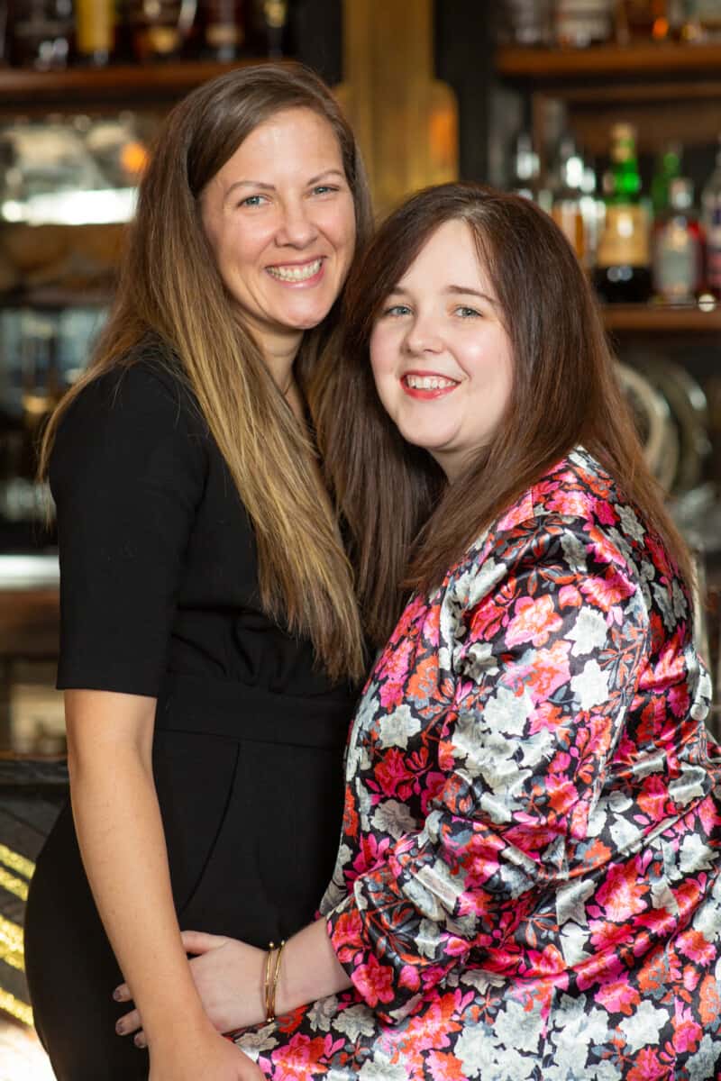 two women, one wearing black jumpsuit, and the other wearing pink flowered jacket, who are LGBTQ couple smile for the camera